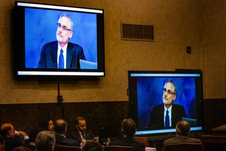 Two screens affixed to a wall show a man testifying in a court case