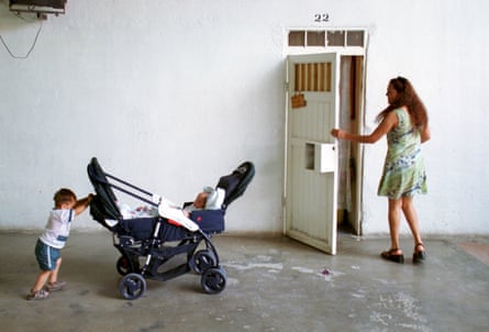 A woman holds a barred door open as a small boy pushes a pram with a baby in it along a corridor