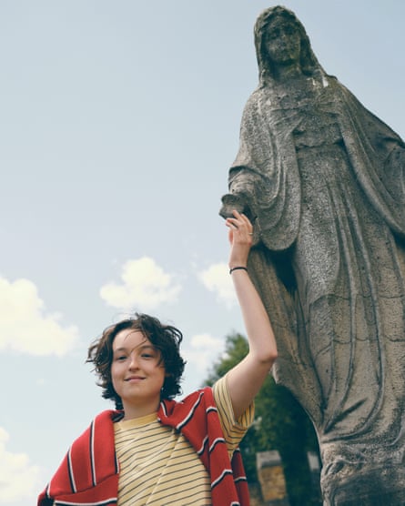 Bella Ramsey wearing a stripy yellow T-shirt with a stripy red jumper over her shoulders holding the hand of a female statue