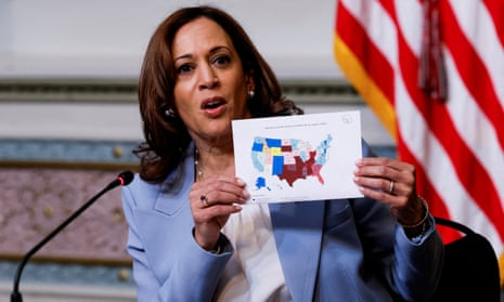 Woman wearing blue suit holds up a map of the United States with a US flag in the background and a microphone in front of her.