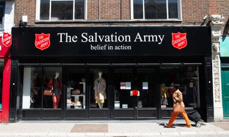 A woman walking past the black shopfront of a Salvation Army shop, which has a a white-lettered signboard with two red shields on either side