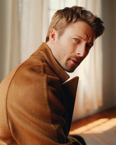 Portrait of Glen Powell against a background of drawn, full-length white curtains wearing a brown coat sitting side-on to the camera and looking to his right at the camera