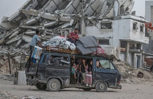 People sit inside a people-carrier with belongings strapped to the roof, amid scenes of wrecked buildings