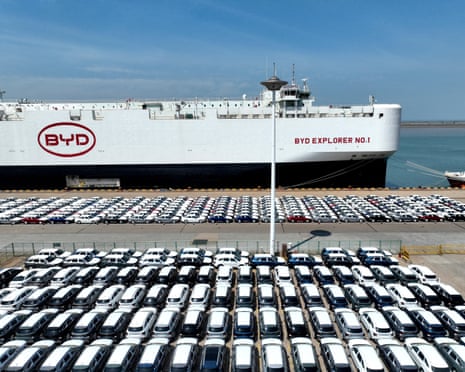 Big number of electric cars parked in foreground with a large ship docked in background