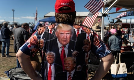 The back of a man wearing a T-shirt with several pictures of Donald Trump, as well as a red baseball hat reading ‘Trump’, at a rally