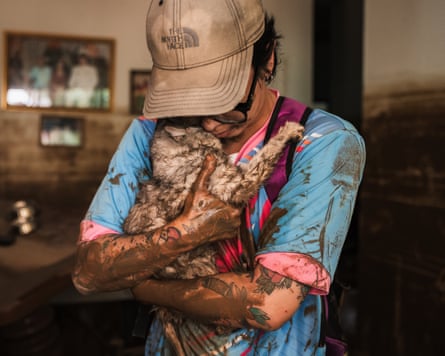 A woman in a baseball cap, wearing black glasses, a blue T-shirt and muddied arms, holds her rescued cat to her face
