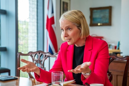 Emma Reynolds sits at a table speaking with her hands held out in front of her. She is in her late 40s with hair in a neat blond bob and wears a bright red jacket over a black top. There is a furled union jack flag by a window behind her.