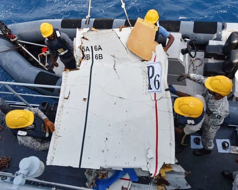 Rescue team members wearing yellow helmets hold part of the cargo plane as it is lifted from a small boat onto a larger vessel