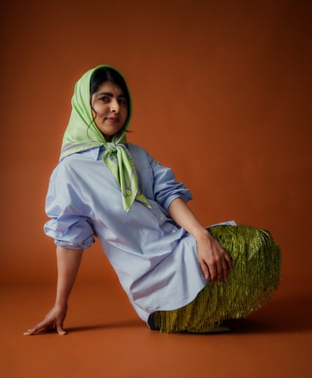 Malala Yousafzai sitting on the floor leaning backwards with her right arm supporting her, wearing a light-blue shirt with rolled-up sleeves, a lim-green textured skirt and a pale green scarf tied underneath her chin