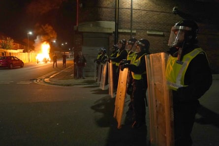 Riot police stand in a line near the entrance to a street where a car has been petrol bombed