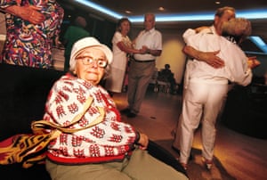 an older woman in a red and white sweater and white hat sits at the edge of the dancefloor smiling while other couples dance