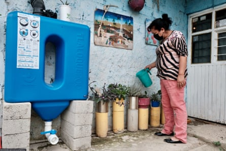 A woman wearing a mask waters pot plants using rainwater collected in a large blue plastic tank.