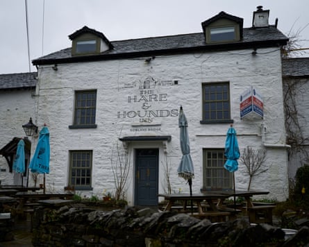 A white historic building called the Hare and Hounds Inn. There are closed blue umbrellas at the front, on dark wooden tables.