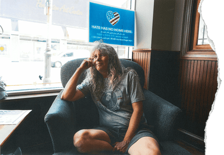 A woman wearing a gray T-shirt and shorts sits on an armchair inside a cafe, a sign reading ‘Hate has no home here’ on the window behind her