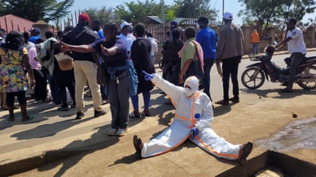 A health worker wearing PPE sits on the ground in the street, with an arm raised in protest. A crowd of people stand behind him.