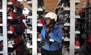 A skier smiles while holding her ski helmet strap amid shelves of boots