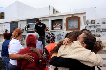 Two women hug, while two others comfort a woman in a red hooded top at a cemetary.