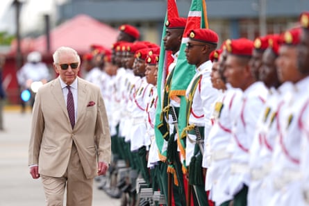 King Charles inspects a guard of honour at the airport as he arrives in Guyana. The military personnel are lined up wearing red hats and white jackets. The king wears a light brown suit and sunglasses.