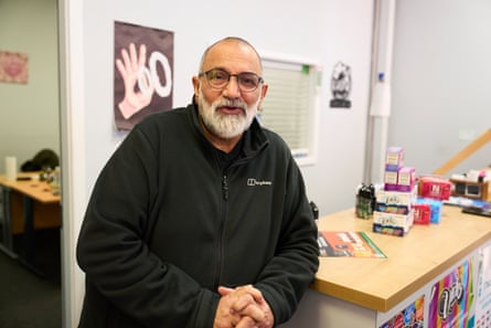 A man with white beard and glasses smiles and leans against a counter on which are several vape products