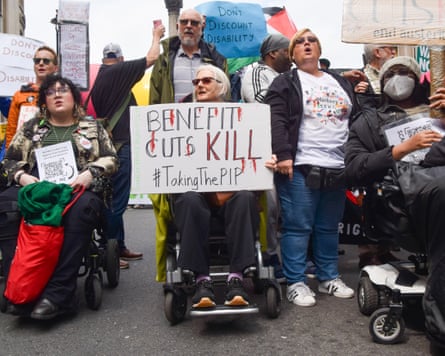 A protester in a wheelchair holds a sign reading: 'Benefit cuts kill'. Around them are other protesters, one holding a sign reading: 'Don't discount disability'.