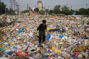 A boy in a long black shirt stands amid a huge pile of rubbish, much of which is plastic bags and packaging