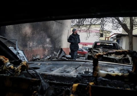 A man looks up to the sky as he walks past a damaged car. The photographer is taking the pic through the glass-less windowscreen of another damaged car