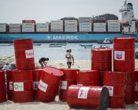 Red oil barrels stacked up on a beach with a container ship in the background
