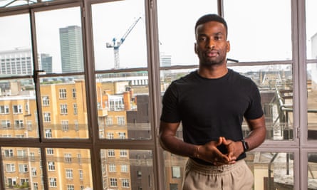 Muyiwa Oki standing looking at the camera, with his back to a large window looking out over London