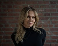 Jennie Garth with blond hair wearing a black top stands before an exposed brick wall.