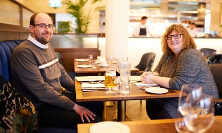 A young, balding man with glasses, wearing a grey top over a white shirt, and an older woman with curly red hair and glasses, wearing a grey sparkly top, sitting at a wooden restaurant table talking