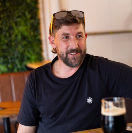 he is sitting in a pub with a pint of what looks like Guinness in front of him. He has short brown hair and a beard, and wears a black T-shirt and has sunglasses balanced on his head; he looks cheerful.