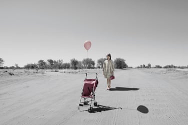 Mother (pram) 2016. A mostly monochrome image of a wide desert with scrubland in the background. In colour, a woman dressed in 60s or 70s style fashion stands holding a pink bag stands some distance apart from a pink stroller with a balloon attached.
