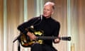 Sting performs sitting on a stool on stage playing a guitar