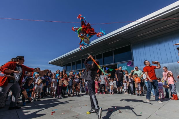 A crowd of people standing in a circle watches a boy standing in the middle with a colorful, paper-covered stick aiming a pinata suspended in the air.