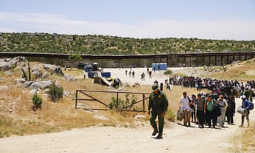 Dozens and dozens of people crowd a dusty path winding up a dry hillside away from a high, rusted, metal wall, led by one man in an olive-green uniform.