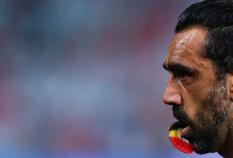 a close up of Adam Goodes holding onto a red, yellow and black mouth guard with his teeth