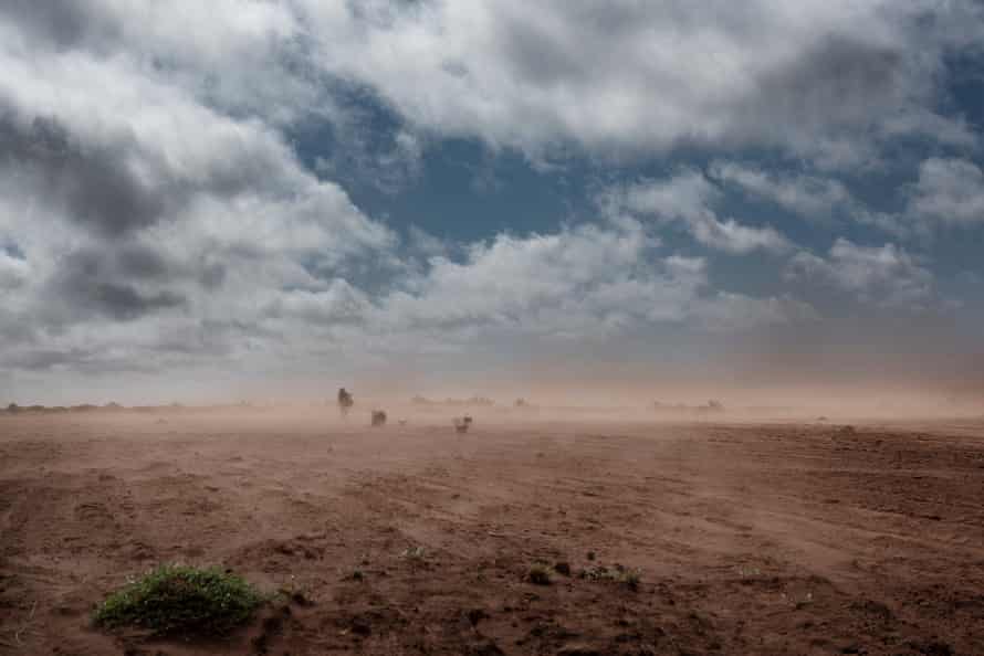 A man hunches against the wind as dust blows across a barren landscape