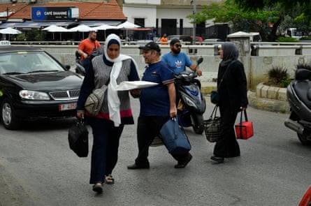 Two women and a man carry bags as they walk through traffic while crossing a road