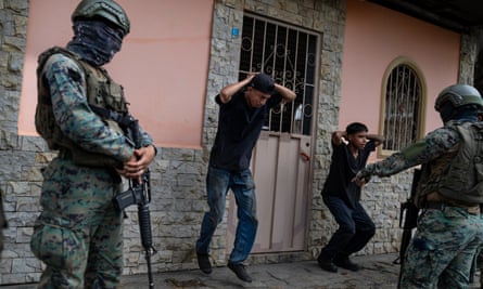 Two young men in black T-shirts exercise next to a pink wall with their hands behind their heads under the direction of two armed soldiers wearing helmets and combat fatigues.
