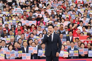 A man in a suit speaking into a microphone in front of a crowd of people holding placards