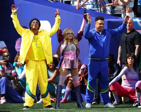 Ronaldinho, Madonna, and Ronaldo in colorful outfits raise their hands on a sports field with a crowd behind them