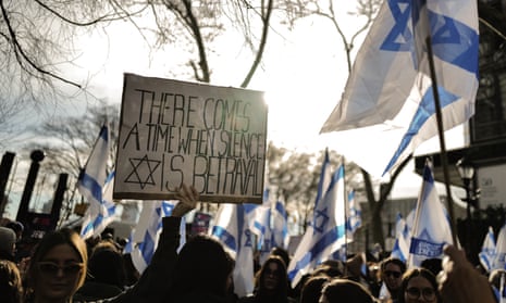 A crowd of demonstrators, pictured from the shoulders up. Many Israeli flags are held aloft, along with a placard in the foreground that reads: 'There comes a time when silence is betrayal.' A wintry sky and bare tree branches are shown behind them.