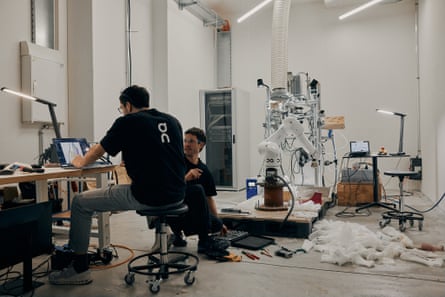 Two men dressed in black work a computer with a pile of soles and shoe material in a studio