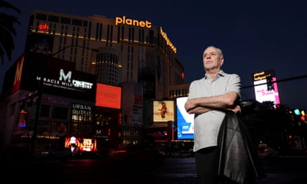 man wearing gray polo shirt and crossing arms stands on street in front of buildings at night