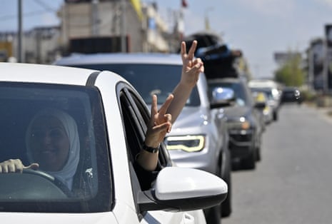 A smiling woman in a white hijab drives a white sedan with one hand and sticks a peace sign out the window with her other. Somebody in her backseat also sticks a peace sidn out the window. Their car is in front of a long line of cars stuck in traffic.
