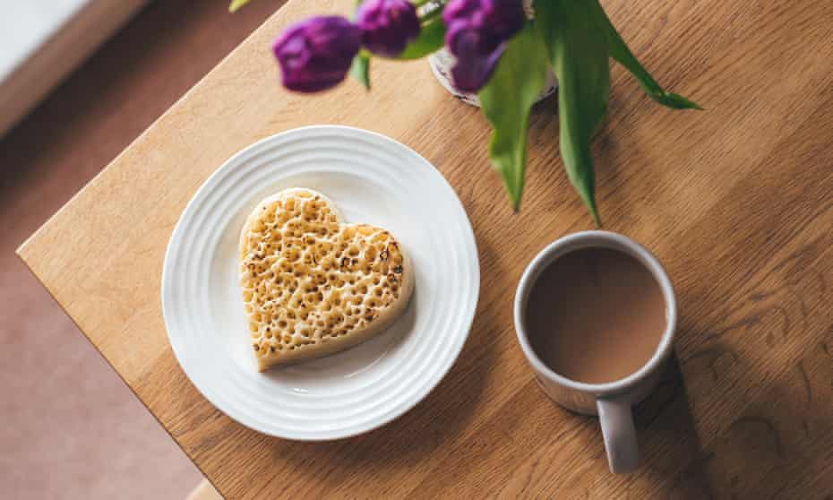 Heart-shaped toasted crumpet on white plate with mug of tea on table with purple tulips
