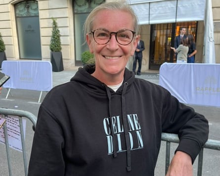 A smiling woman with glasses and her hair tied back, leaning against a metal crowd barrier outdoors. She is wearing a black pullover hoodie with Celine Dion printed on the front