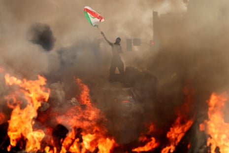 A man waves a flag as smoke and flames rise from a burning vehicle during a protest against federal immigration sweeps, in downtown Los Angeles, California, US, 8 June, 2025.