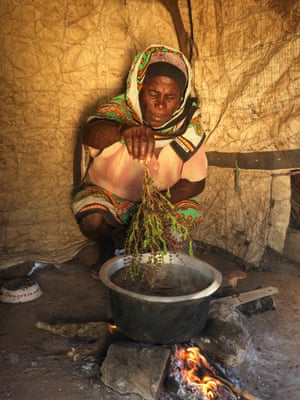An old woman puts a sprig into a bowl of water on an open fire