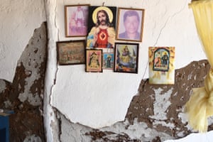 Religious images and two family photographs hang on a cracked interior wall with missing chunks of plaster
